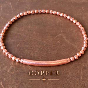 Könnte beinhalten: Ein Armband aus kleinen, runden, metallisch kupferfarbenen Perlen in kreisförmiger Anordnung, mit einer einzelnen, längeren, gebogenen, zylindrischen, kupferfarbenen Röhrenperle in der unteren Mitte. Das Wort COPPER steht darunter.