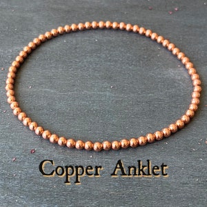 Peut inclure: Un bracelet de cheville en cuivre composé de petites perles rondes. Le bracelet est sur un fond gris.