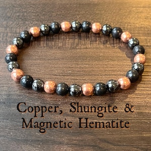 Może przedstawiać: Bransoletka z naprzemiennymi miedzianymi i czarnymi koralikami. Bransoletka wykonana jest z miedzi, szungitu i magnetycznego hematytu. Tekst "Copper, Shungite & Magnetic Hematite" jest wydrukowany poniżej.