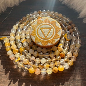 May include: A beaded necklace with a wooden pendant. The necklace features round, translucent beads in shades of yellow and amber. The pendant is circular, with a carved floral design and a central triangle.