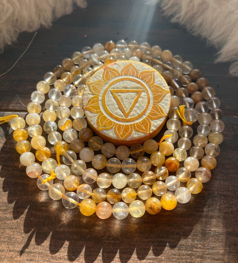 May include: A beaded necklace with a wooden pendant. The necklace features round, translucent beads in shades of yellow and amber. The pendant is circular, with a carved floral design and a central triangle.