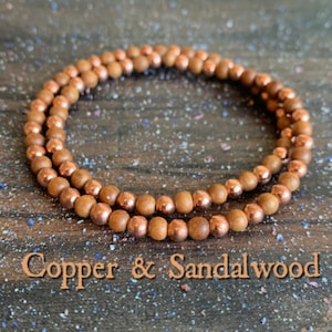 Peut inclure: Un bracelet à double rang de perles en cuivre et en bois de santal. Les perles sont d'une couleur brun chaud et les perles en cuivre ont une finition brillante. Le bracelet est sur une surface en bois.