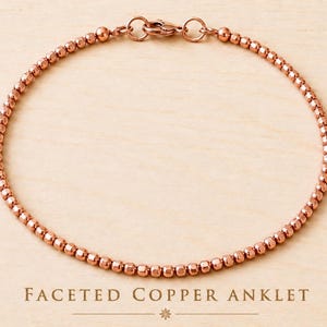 May include: A copper anklet composed of small, faceted, metallic beads arranged in a continuous circular strand. The anklet features a secure lobster claw clasp mechanism at the top. The text "FACETED COPPER ANKLET" is printed below the jewelry.