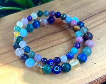 May include: Two beaded bracelets with a variety of colourful gemstones, including blue, green, purple, and pink. One bracelet features an evil eye bead. The bracelets are displayed on a wooden surface.