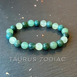 May include: A green and light green gemstone bracelet with the text "Taurus Zodiac" on a dark gray surface.