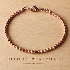 May include: A bracelet composed of small, metallic, rose-gold coloured faceted beads arranged in a continuous circular strand. The bracelet features a secure lobster claw clasp mechanism at the top, connecting the two ends of the beaded chain.
