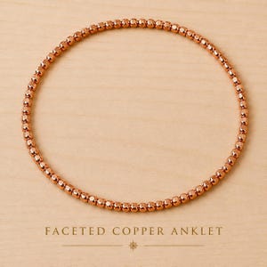 Peut inclure: Une chevillère circulaire composée de petites perles métalliques de couleur cuivre avec une finition facettée. Les perles sont enfilées en une boucle continue, créant une surface réfléchissante qui capte la lumière. Le texte FACETED COPPER ANKLET est en dessous.