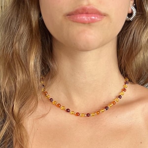 May include: A beaded necklace featuring small, round, polished amber-colored beads in varying shades of golden yellow, orange, and deep reddish-brown. The beads are strung together with small, reflective silver-toned spacers between each individual bead.