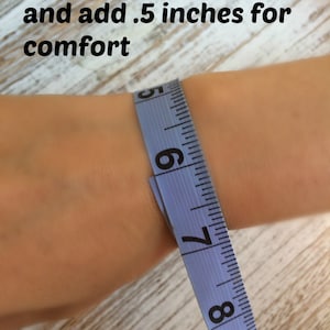 May include: A light blue measuring tape wrapped around a wrist, showing the measurement near the 6-inch mark. The text "Measure your wrist and add .5 inches for comfort" is printed in black over the image to provide sizing instructions.