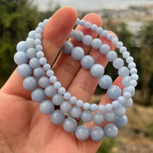 Puede incluir: Tres pulseras de cuentas azul claro con cuentas de diferentes tama&ntilde;os. Las pulseras est&aacute;n hechas de cuentas lisas y redondas y est&aacute;n dispuestas en capas. Se sostienen en una mano, con un fondo natural borroso.