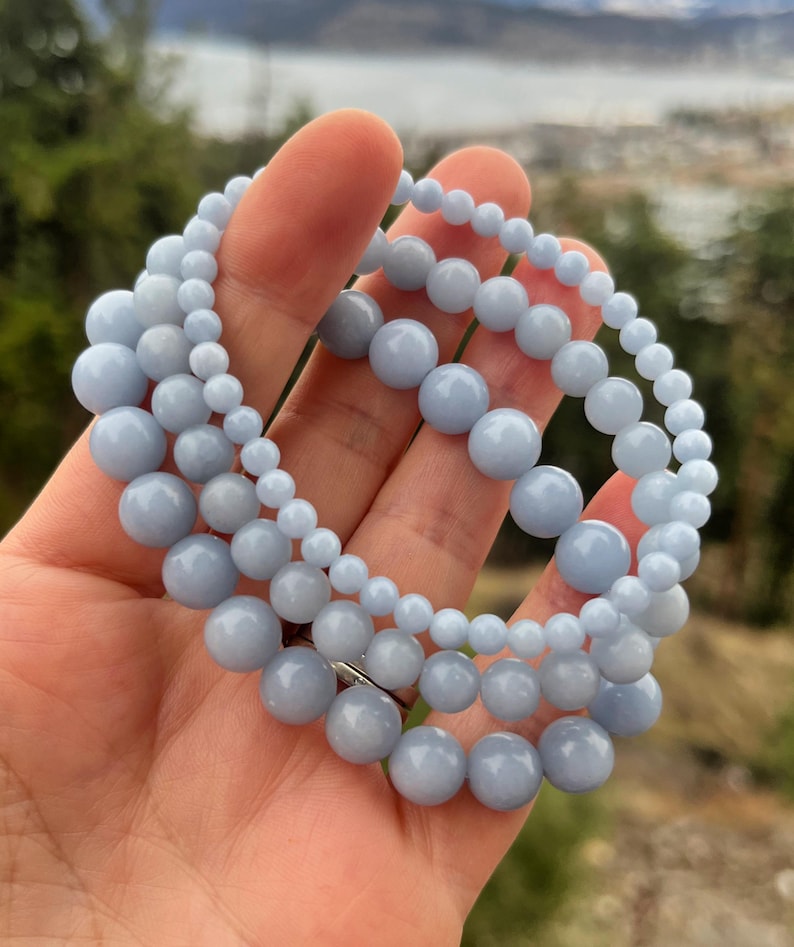 Puede incluir: Tres pulseras de cuentas azul claro con cuentas de diferentes tama&ntilde;os. Las pulseras est&aacute;n hechas de cuentas lisas y redondas y est&aacute;n dispuestas en capas. Se sostienen en una mano, con un fondo natural borroso.