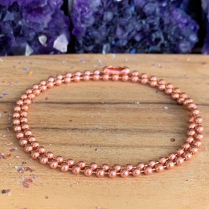 May include: A copper chain necklace with a double row of small, round beads. The necklace is on a wooden surface.