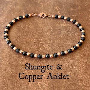 Puede incluir: Una tobillera con cuentas negras y color cobre alternadas. La joya está hecha de pequeñas cuentas redondas ensartadas con un cierre de cobre. El texto "Shungite & Copper Anklet" está impreso debajo.