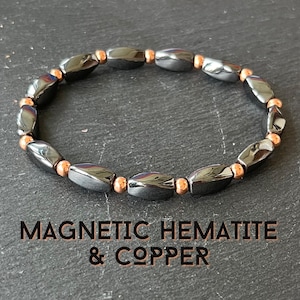 May include: A bracelet made of polished, dark gray magnetic hematite beads and copper-colored beads. The bracelet is strung together, forming a complete circle. The text "MAGNETIC HEMATITE & COPPER" is displayed below the bracelet.