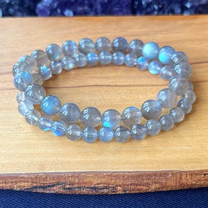 May include: A double-strand bracelet made of iridescent grey labradorite beads. The bracelet is on a wooden surface.