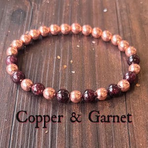 Könnte beinhalten: Ein Armband aus Kupfer- und Granatperlen. Das Armband wechselt zwischen kupferfarbenen und tiefroten Granatperlen. Der Text "Copper & Garnet" wird darunter angezeigt.