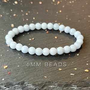 Puede incluir: Una pulsera de cuentas azul claro con cuentas de 6 mm. La pulsera est&aacute; hecha de cuentas redondas y lisas y se muestra sobre una superficie oscura y texturizada. El texto "6MM BEADS" es visible debajo.