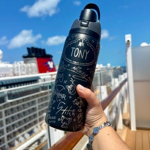 Puede incluir: Una botella de agua negra mate con tapa negra y asa de transporte. La botella presenta dibujos de líneas blancas de varios personajes y el texto TONY, WHERE DREAMS COME TRUE, MICKEY, STITCH y LIGHT, junto con pequeñas estrellas y corazones.