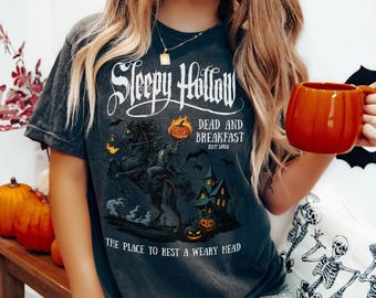 May include: Dark gray t-shirt with a Halloween-themed graphic. The design features the words "Sleepy Hollow Dead and Breakfast" with a headless horseman, a haunted house, and pumpkins. The text "The Place to Rest a Weary Head" is also included.