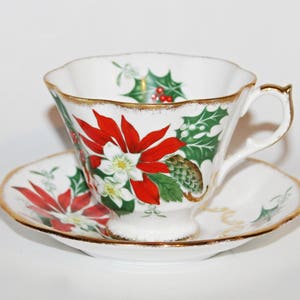 May include: A white teacup and saucer set with a gold trim, decorated with red poinsettias and green holly. The teacup and saucer have a festive design, perfect for the holiday season. A classic tea set.