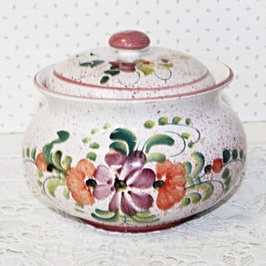 May include: A ceramic jar with a lid, decorated with hand-painted flowers in orange, purple, and green. The jar has a speckled white finish and a pink rim and knob. A decorative kitchen item.