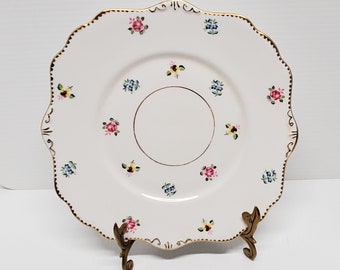 May include: A white, scalloped-edge plate with a gold rim and floral pattern. The plate features small clusters of pink, blue, and yellow flowers. The plate is displayed on a decorative gold stand.