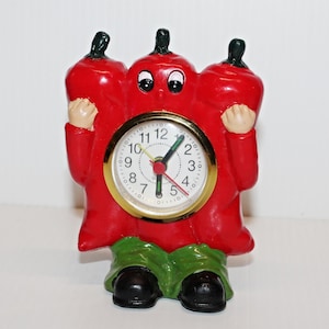 May include: A novelty clock in the shape of a red chili pepper, featuring a gold-coloured clock face. The clock has black eyes, small tan hands, green trousers, and black shoes. The clock face shows the time.