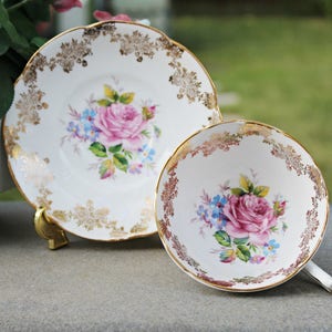 May include: A vintage teacup and saucer set. The white porcelain features a pink rose and blue floral design in the center, with gold trim and accents. The saucer and cup are displayed on a gray surface, with a green background.