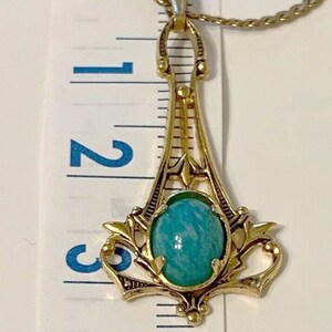 May include: Gold-toned pendant necklace featuring a turquoise-colored oval stone. The pendant has an ornate, Art Deco-inspired design with a decorative frame. The necklace is approximately 3 inches in length.