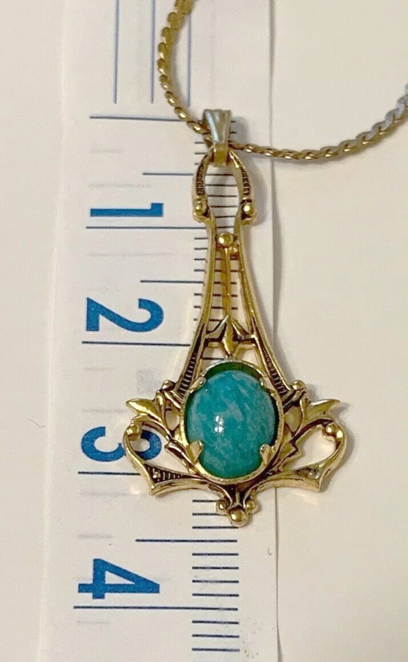 May include: Gold-toned pendant necklace featuring a turquoise-colored oval stone. The pendant has an ornate, Art Deco-inspired design with a decorative frame. The necklace is approximately 3 inches in length.