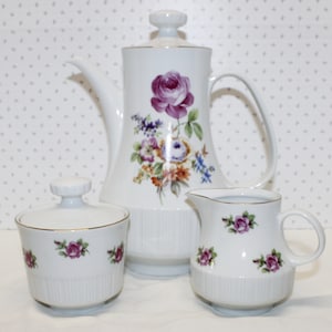 May include: A white porcelain tea set with a floral design. The set includes a teapot, creamer, and sugar bowl. The teapot has a lid and a handle. The creamer and sugar bowl have lids.