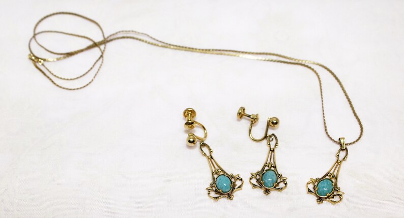 May include: A vintage jewelry set featuring a gold-toned necklace and matching earrings. The necklace and earrings have a filigree design with a turquoise-colored stone in the center. The earrings are clip-on style.
