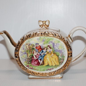 Peut inclure: Théière en céramique crème avec garniture et anse dorées. Le panneau central ovale représente des personnages en costume d'époque, encadré d'une bordure dorée. Le bec et le couvercle sont également dorés. Objet décoratif.