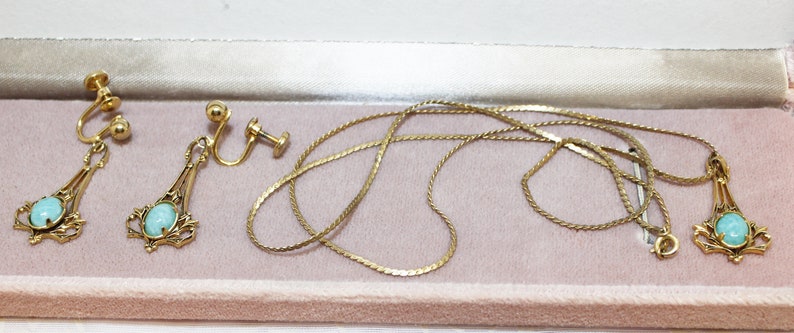 May include: A vintage jewelry set featuring gold-toned metal. The set includes a necklace with a pendant and a pair of clip-on earrings. Each piece has a light blue oval stone. The jewelry is displayed in a pink velvet-lined box.