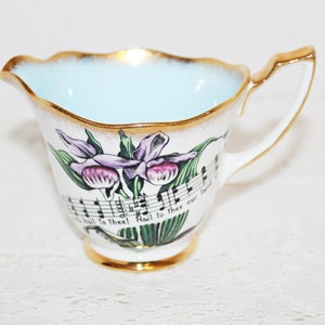 May include: A white porcelain creamer with a light blue interior and gold trim. The creamer features a floral design with purple flowers and green leaves, and the words "Hail to thee! Hail to thee our Lord" are printed on the side.
