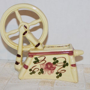 May include: A ceramic spinning wheel planter with a white base, pink and brown accents, and a green floral design. The wheel is painted in brown and white.