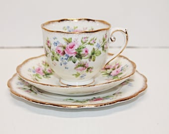 May include: A white teacup and saucer set with a floral pattern of pink roses and blue forget-me-nots. The set has a gold trim around the edges.