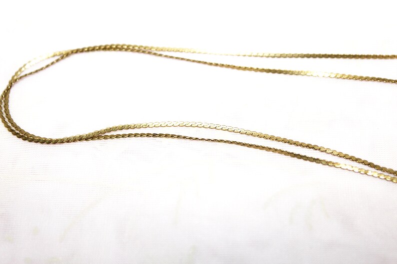 May include: A close-up shot of two delicate gold necklaces. The necklaces are made of thin, flat, circular links that catch the light. The chains are displayed against a white background, highlighting their intricate design and warm color.