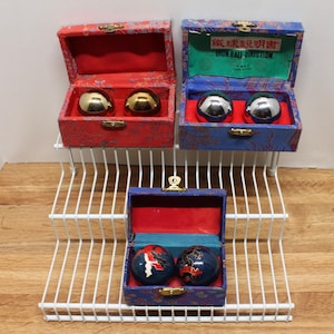 May include: Three sets of Chinese health balls in decorative boxes. The boxes are red, blue, and patterned with gold clasps. The balls are gold, silver, and blue with red and white designs. The boxes are displayed on a white wire shelf.