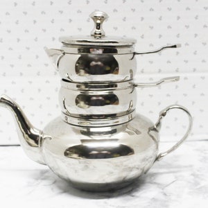 May include: A shiny, silver-colored tea set with a teapot and two stacked infusers. The teapot has a curved spout and a handle. The infusers have small handles. The set is made of metal and sits on a white surface.