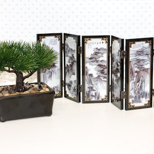 May include: A miniature artificial pine bonsai tree in a rectangular black ceramic pot filled with small stones, positioned next to a six-panel folding screen featuring black and white landscape ink wash paintings with gold geometric borders.
