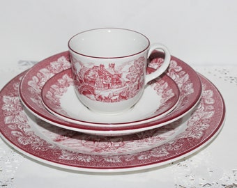 May include: A white teacup and saucer set with a red transferware design featuring a rural landscape scene. The set includes a cup, a small plate, and a larger plate. The plates have a floral border.