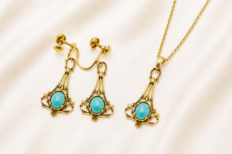 May include: A gold-toned jewelry set featuring a necklace and clip-on earrings. Each piece has a filigree design and a central oval turquoise stone. The set is displayed on a white, textured surface.