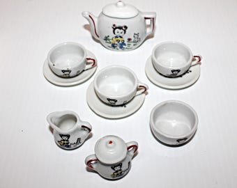 May include: A miniature white ceramic tea set featuring a teapot, cups, saucers, a creamer, and a sugar bowl. Each piece is decorated with a cartoon bear design and red trim. The set is arranged on a white surface.