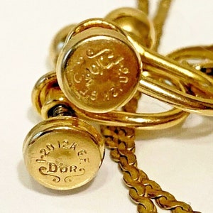 May include: Close-up of gold-toned jewelry, possibly earrings or a necklace. The pieces have a shiny finish and feature engraved text, including the word "Dior". The design includes a chain and screw-like details.