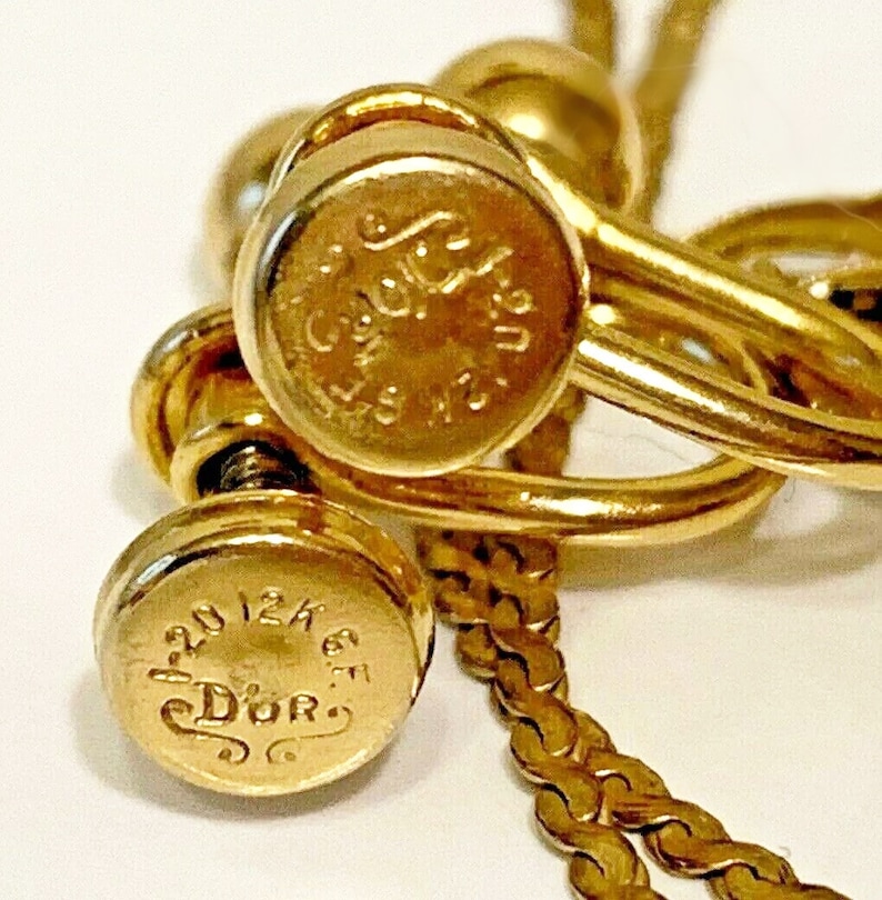 May include: Close-up of gold-toned jewelry, possibly earrings or a necklace. The pieces have a shiny finish and feature engraved text, including the word "Dior". The design includes a chain and screw-like details.