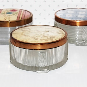 May include: Three vintage glass jars with copper lids and floral patterned tops. The jars are clear glass with a ribbed design. The lids are a shiny copper colour. The tops are made of fabric with floral patterns in pink, blue, and white.