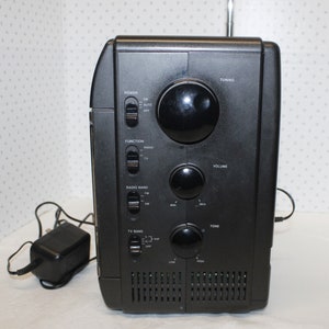 May include: Black radio with a silver antenna, multiple knobs and buttons, and a power cord. The radio has labels for "Power", "Function", "Radio Band", "TV Band", "Tuning", "Volume", and "Tone".
