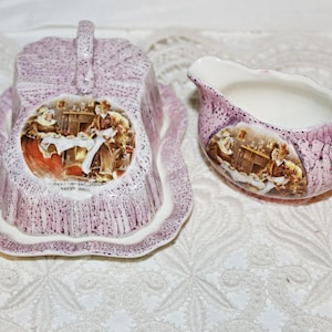 May include: A vintage purple and white ceramic butter dish and creamer set with a floral design and a scene of people gathered around a table. The set features a handle on the butter dish and a spout on the creamer.