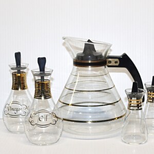 May include: A set of four glass bottles with gold trim and black stoppers. Two bottles are labelled "Vinegar" and "Oil". The bottles are on a white surface.
